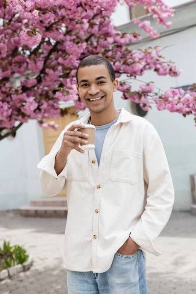 Giovane uomo africano americano che tiene bevanda da asporto vicino all'albero di ciliegio rosa sulla strada — Foto stock