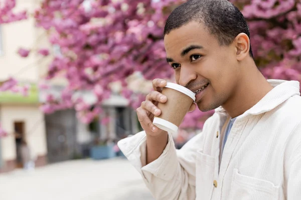Happy african american man drinking coffee to go near pink cherry tree — Stock Photo
