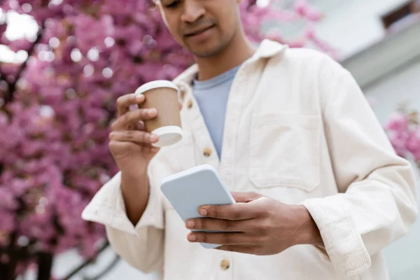 Vista ritagliata di uomo afroamericano in possesso di caffè per andare e utilizzando smartphone vicino all'albero di ciliegio rosa — Foto stock