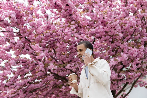 Young african american man holding takeaway drink and talking on smartphone near pink cherry tree — Stock Photo