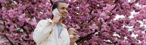 Smiling african american man holding takeaway drink and talking on smartphone near blooming cherry tree, banner — Stock Photo