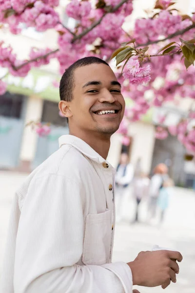 Positiver afrikanisch-amerikanischer Mann lächelt in der Nähe blühender Kirschbäume — Stockfoto