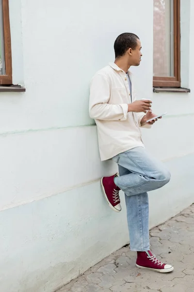 Junger afrikanisch-amerikanischer Mann hält Pappbecher und Smartphone in der Hand, während er sich an ein Gebäude auf der Straße in Europa lehnt — Stockfoto