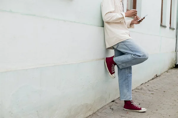 Cropped view of african american man holding paper cup and smartphone while leaning on building on street in europe — Stock Photo