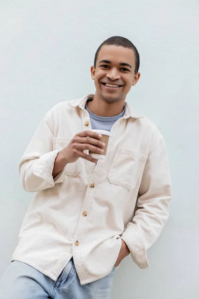 Feliz joven afroamericano hombre sosteniendo taza de papel cerca de edificio en la calle en Europa - foto de stock