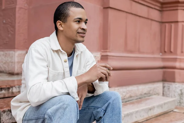 Junger afrikanisch-amerikanischer Mann hält Imbissgetränk in der Hand und sitzt in der Nähe eines Gebäudes auf einer Straße in Europa — Stockfoto