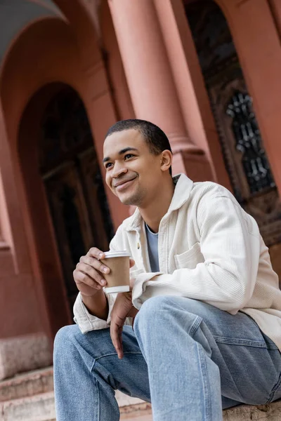Glücklicher junger afrikanisch-amerikanischer Mann hält Pappbecher in der Hand und sitzt in der Nähe eines Gebäudes auf der Straße in Europa — Stockfoto