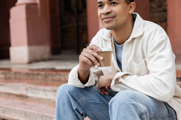 Heureux homme afro-américain tenant boisson à emporter et assis près du bâtiment dans la rue en Europe — Photo de stock