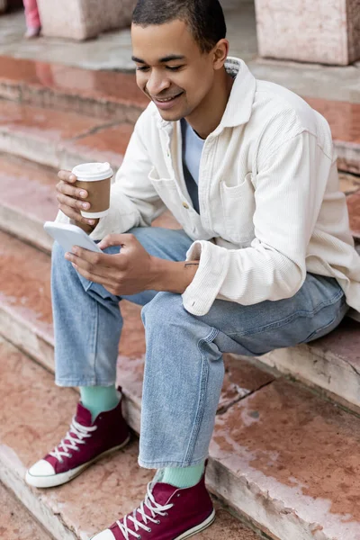 Hombre afroamericano feliz sosteniendo una bebida para llevar y usando un teléfono inteligente mientras está sentado en las escaleras de la ciudad - foto de stock