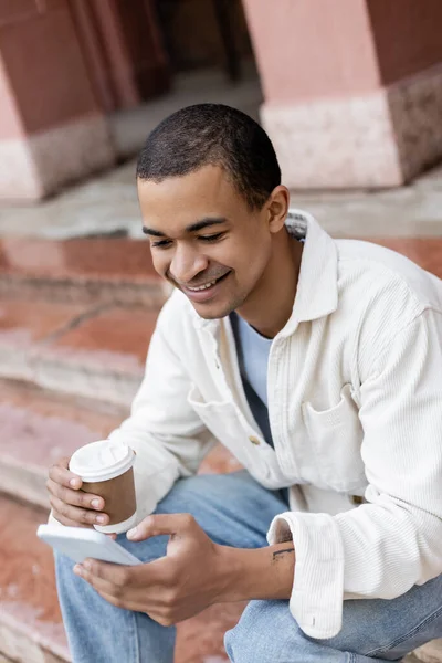 Alegre afroamericano hombre celebración takeaway bebida y el uso de teléfono inteligente mientras se sienta en escaleras en la ciudad - foto de stock