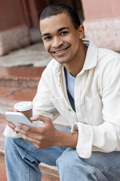 Heureux homme afro-américain tenant boisson à emporter et smartphone tout en étant assis sur les escaliers en ville — Photo de stock