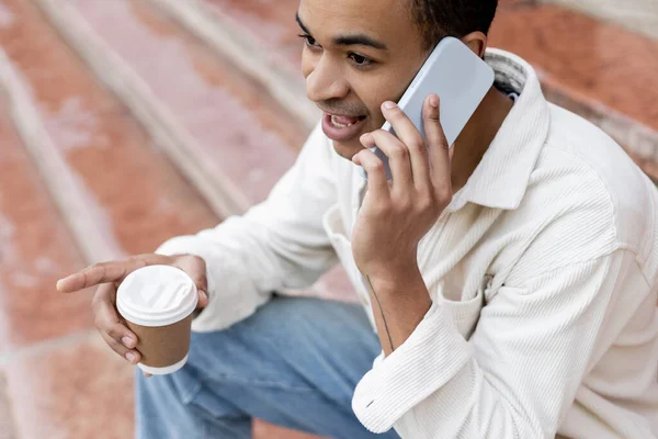 African american man holding takeaway beverage and talking on smartphone while sitting on stairs in city — Stock Photo