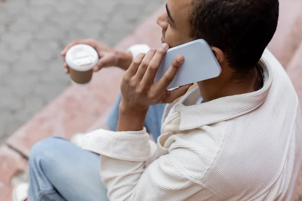 Top view of african american man holding takeaway beverage and talking on smartphone while sitting on stairs in city — Stock Photo