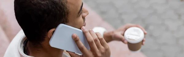 Vista superior del hombre afroamericano sosteniendo una bebida para llevar y hablando en un teléfono inteligente, pancarta - foto de stock