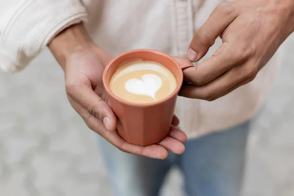 Vista cortada do homem afro-americano segurando takeaway bebida de café com arte latte na xícara — Fotografia de Stock