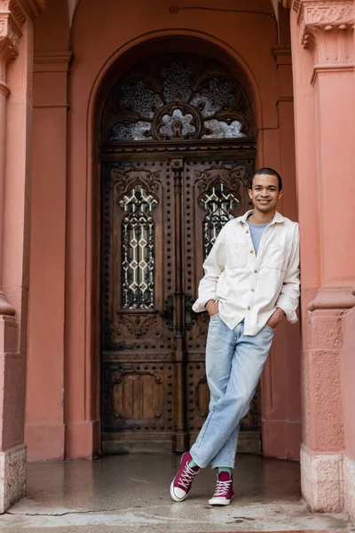 Longitud completa de hombre afroamericano feliz en camisa chaqueta posando con las manos en bolsillos cerca de la construcción en Europa - foto de stock