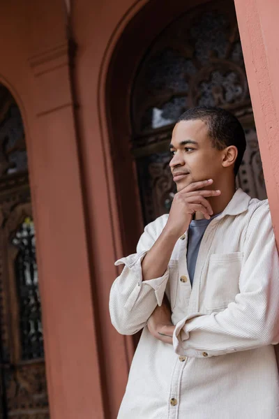 Hombre afroamericano pensativo en camisa chaqueta posando cerca de edificio en Europa - foto de stock