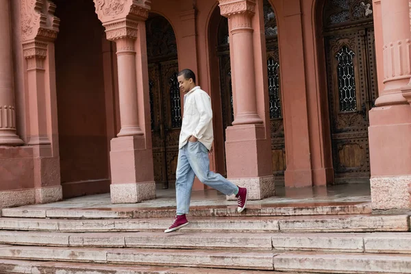 Tutta la lunghezza dell'uomo afroamericano in camicia giacca camminare con le mani in tasche vicino edificio in Europa — Foto stock