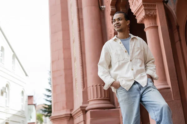 Visão de baixo ângulo do homem americano africano satisfeito em camisa jaqueta de pé com as mãos em bolsos perto do edifício — Fotografia de Stock