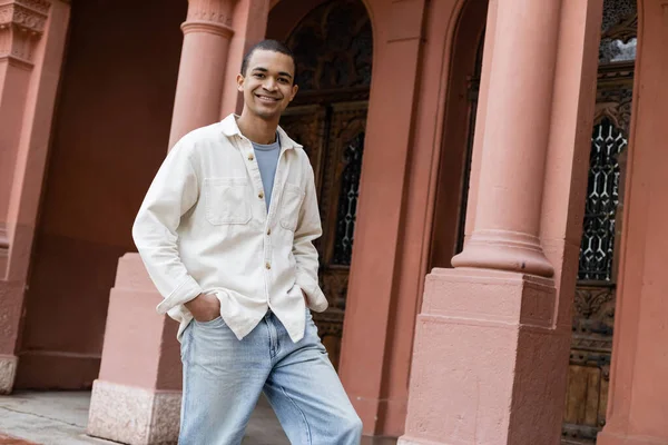 Joyeux homme afro-américain veste chemise debout avec les mains dans les poches près du bâtiment — Photo de stock