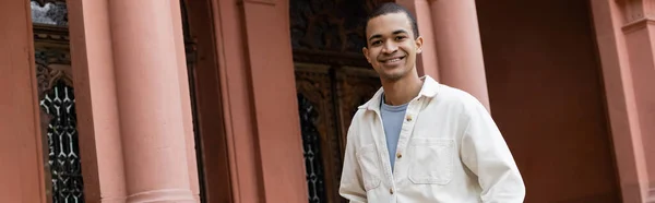 Joyful african american man in shirt jacket standing near building, banner — Stock Photo