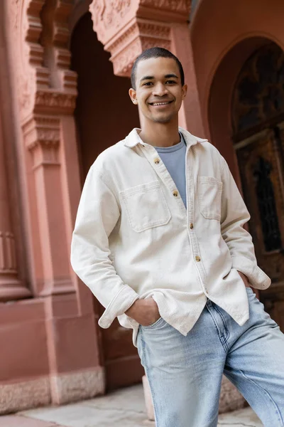 Stylish african american man in shirt jacket standing with hands in pockets and smiling near building — Stock Photo
