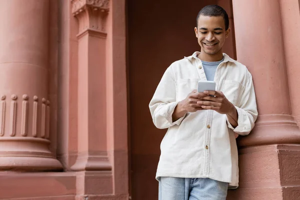 Joyful african american man in shirt jacket messaging on smartphone near building — Stock Photo