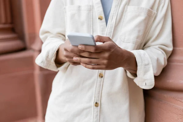 Vista ritagliata dell'uomo afroamericano in camicia giacca messaggistica su smartphone vicino al palazzo — Foto stock