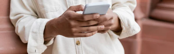 Cropped view of african american man in shirt jacket messaging on smartphone near building, banner — Stock Photo