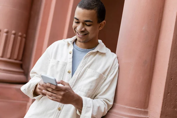 Радісний афроамериканський чоловік у сорочці піджак смс на смартфон біля будівлі — стокове фото