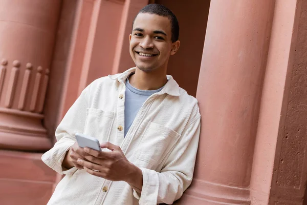Joyful african american man in shirt jacket holding smartphone near building — Stock Photo