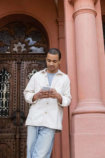 Young african american man in shirt jacket chatting on smartphone near building — Stock Photo
