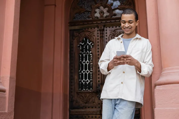 Happy african american man in shirt jacket chatting on smartphone near building — Stock Photo