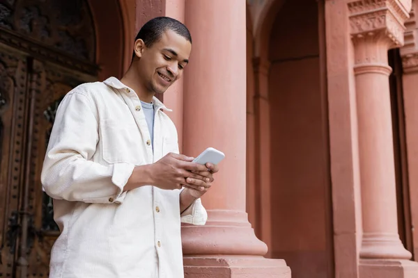 Cheerful african american man in stylish shirt jacket messaging on smartphone near building — Stock Photo