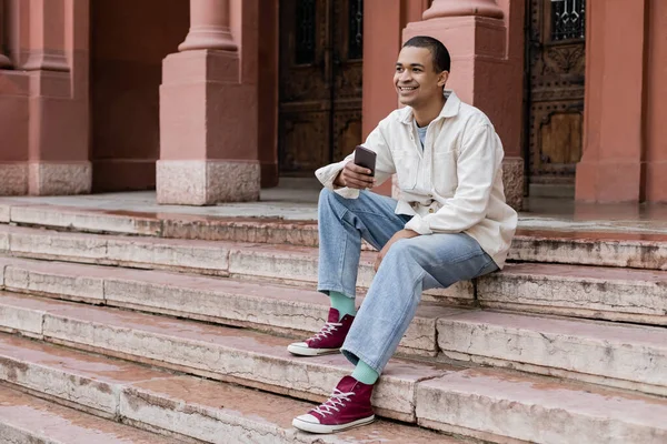 Pleine longueur de heureux homme afro-américain tenant smartphone tout en étant assis sur les escaliers en ville — Photo de stock