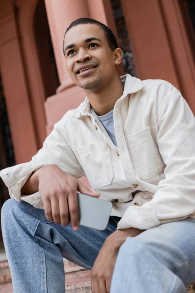 Vista de ángulo bajo del hombre afroamericano feliz en chaqueta de camisa que sostiene el teléfono móvil en la ciudad - foto de stock