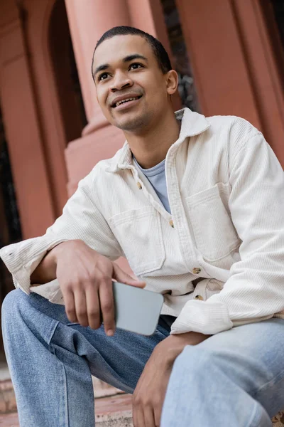 Low angle view of happy african american man in shirt jacket holding mobile phone in city — Stock Photo