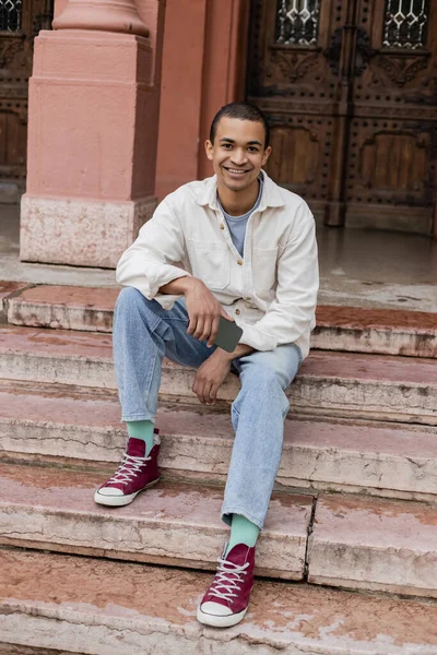 Полная длина довольный африканский американец в рубашке куртке держа мобильный телефон, сидя на лестнице в городе — стоковое фото