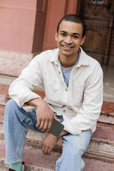 Joyful african american man in shirt jacket holding mobile phone while sitting on stairs in city — Stock Photo