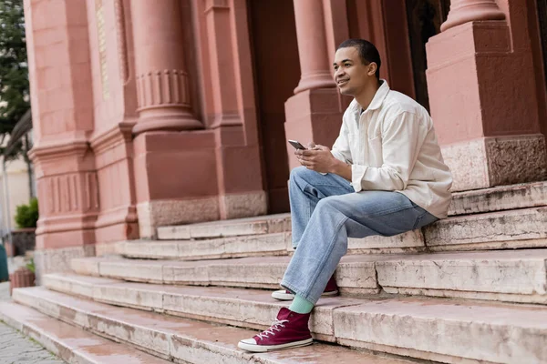 Full length of happy african american man in shirt jacket holding mobile phone while sitting on stairs in city — Stock Photo