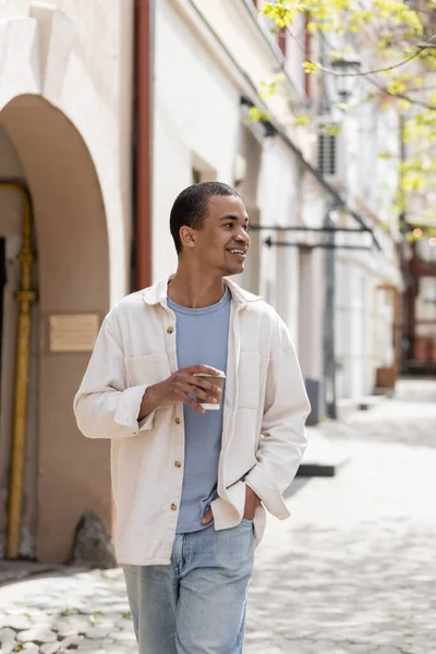 Hombre afroamericano feliz en chaqueta de camisa sosteniendo bebida para llevar y caminando con la mano en el bolsillo en la ciudad urbana - foto de stock