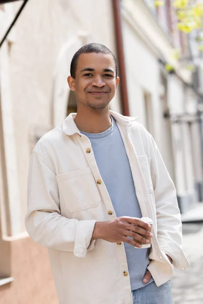 Sorridente afro-americano homem de camisa jaqueta segurando takeaway bebida na cidade urbana — Fotografia de Stock