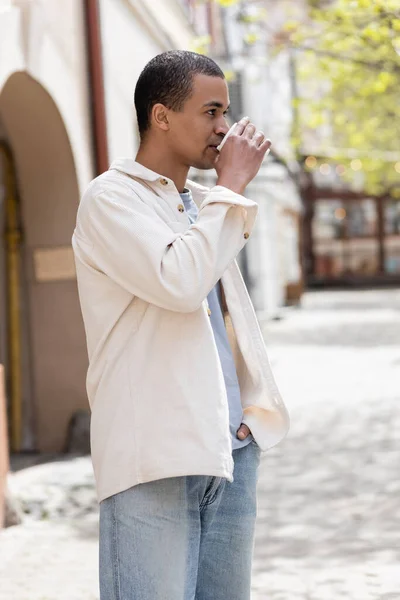 Young african american man in shirt jacket drinking takeaway beverage and standing with hand in pocket in urban city — Stock Photo