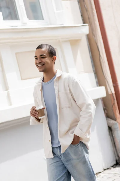 Cheerful african american man in shirt jacket holding coffee to go and walking in urban city — Stock Photo