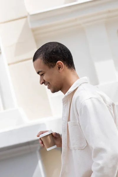 Vista lateral del hombre afroamericano feliz en camisa chaqueta sosteniendo café para ir y caminar en la ciudad urbana - foto de stock