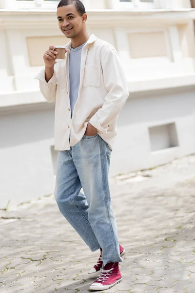 Full length of happy african american man in shirt jacket holding coffee to go and walking in urban city — Stock Photo