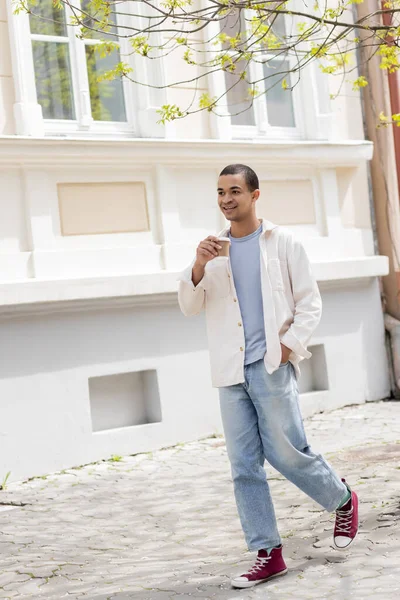Pleine longueur de heureux homme afro-américain en veste chemise et jeans tenant café pour aller et marcher dans la ville urbaine — Photo de stock