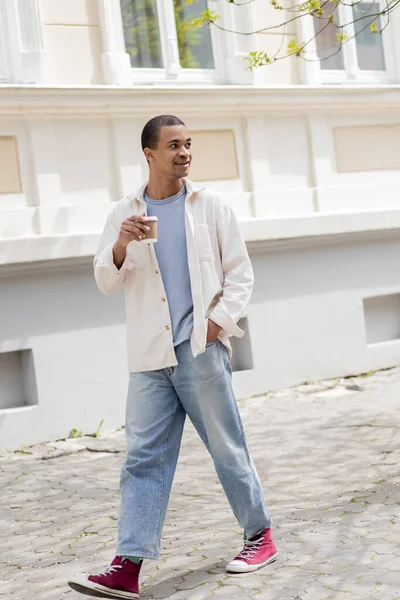 Comprimento total de homem americano africano positivo em camisa casaco e jeans segurando café para ir e caminhar na cidade urbana — Fotografia de Stock