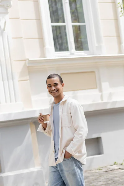 Задоволений афроамериканський чоловік у сорочці і джинсах тримає каву, щоб піти і ходити в міському місті — стокове фото