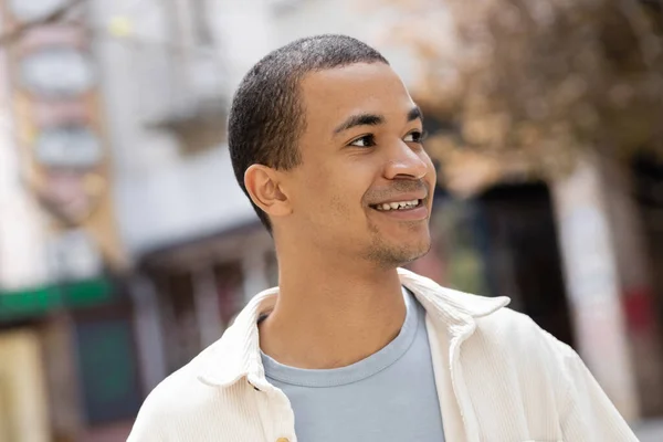 Молодой и улыбающийся африканский американец в рубашке смотрит в сторону — стоковое фото